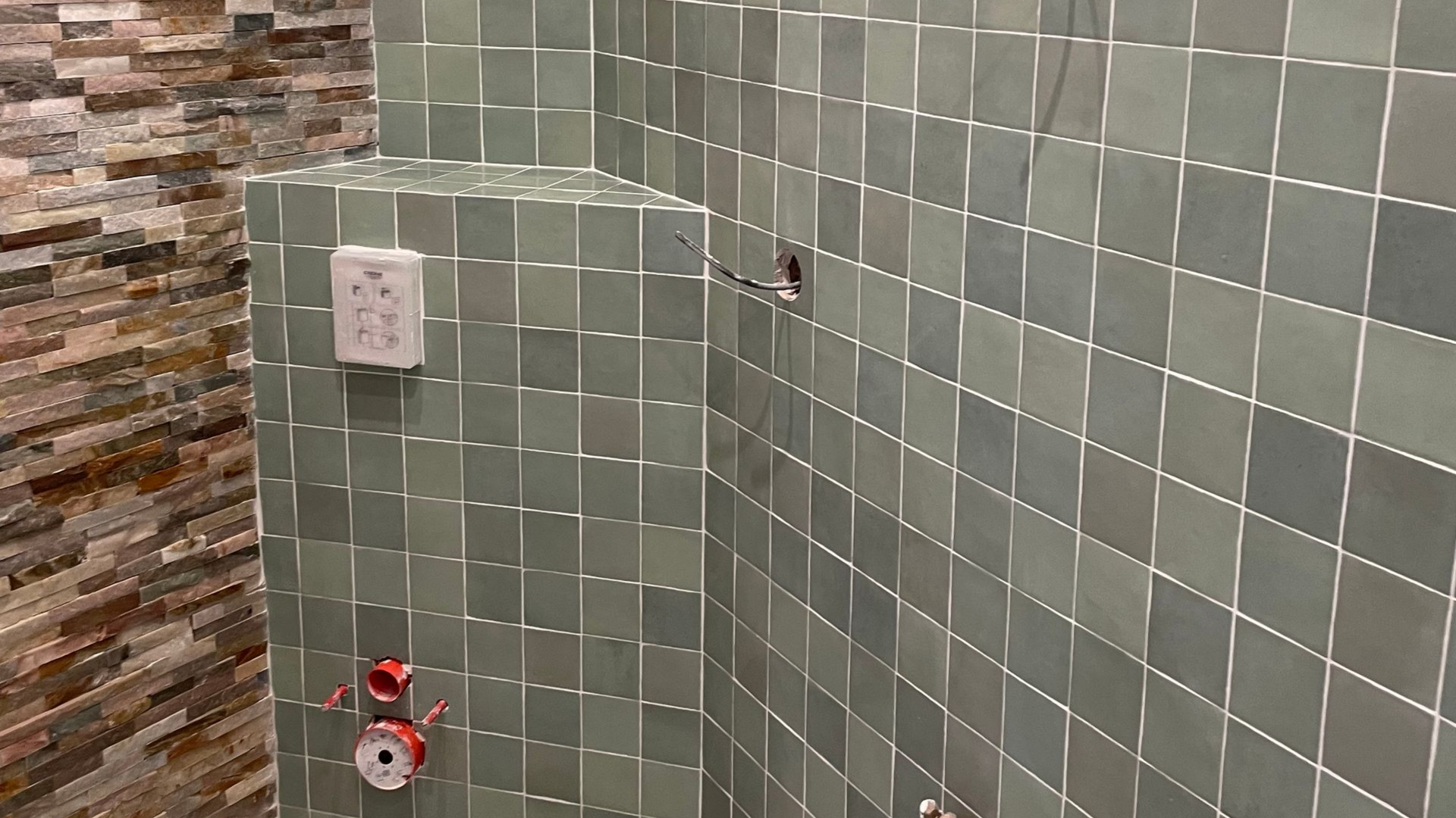 Douche carrelée verte avec prise électrique et tuyauterie apparente.