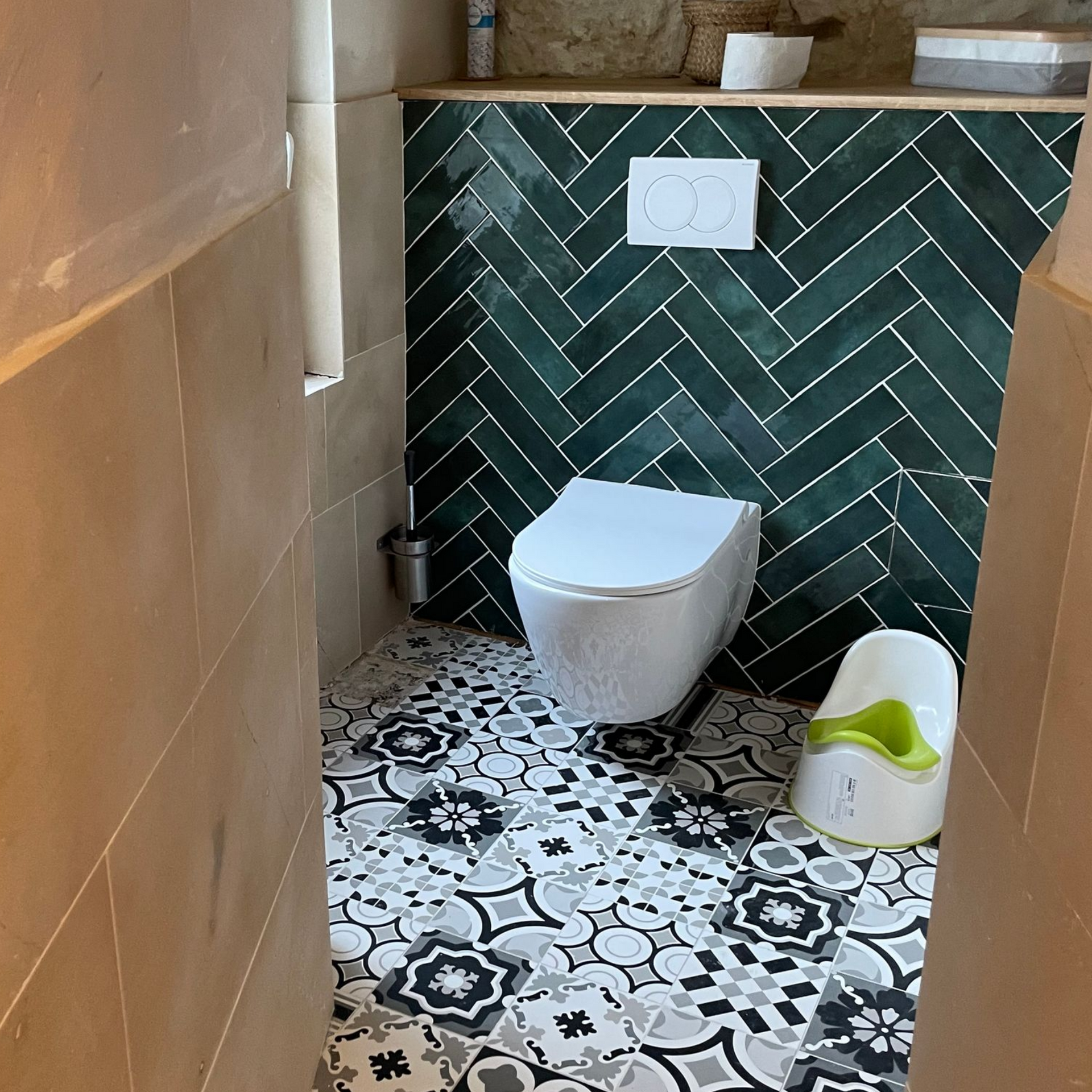 Petite salle de bains avec carrelage vert à chevrons, sol à motifs, toilettes blanches et siège de toilette.