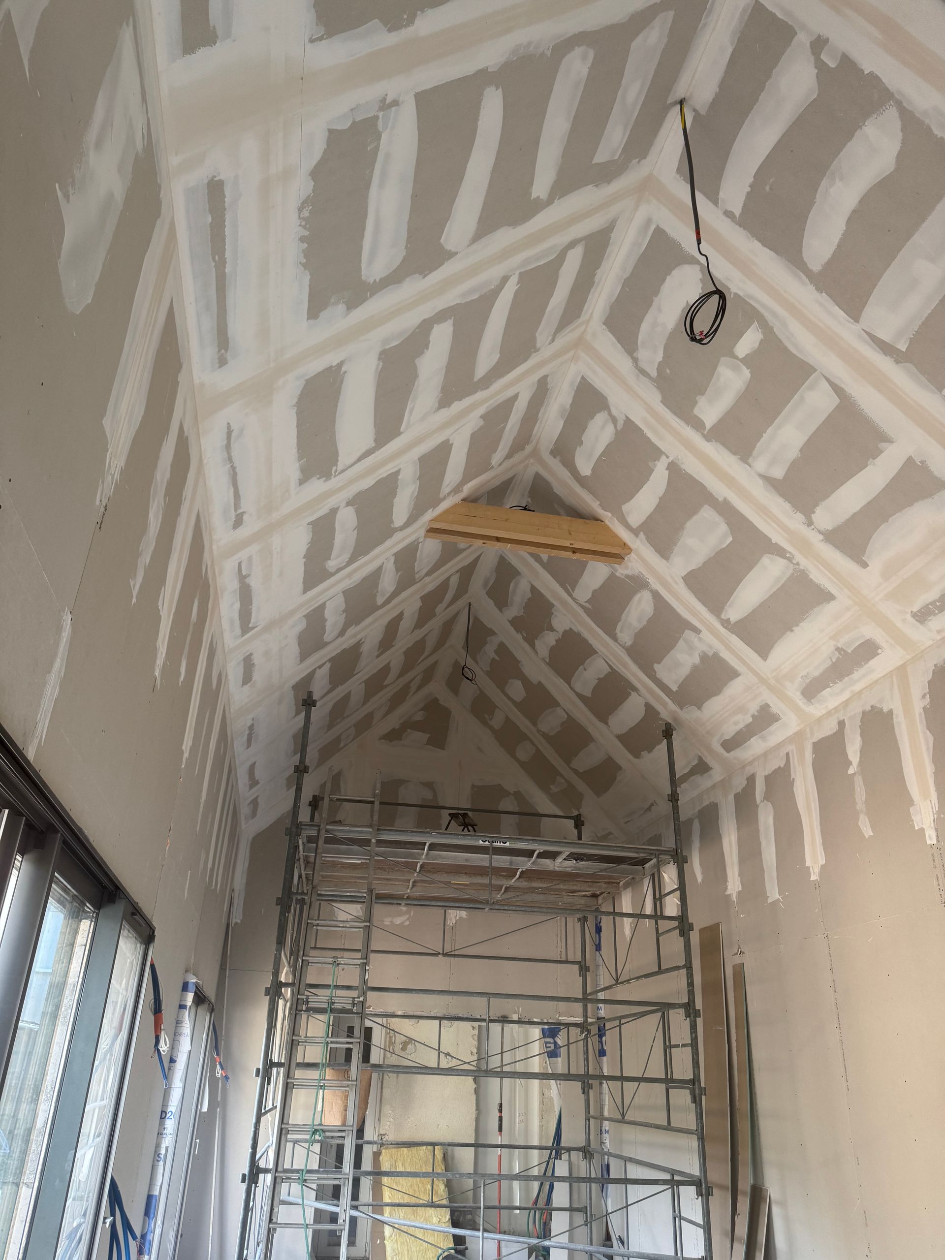 Vue intérieure d'une pièce en construction avec un plafond haut et incliné ; cloisons sèches installées, échafaudages présents.