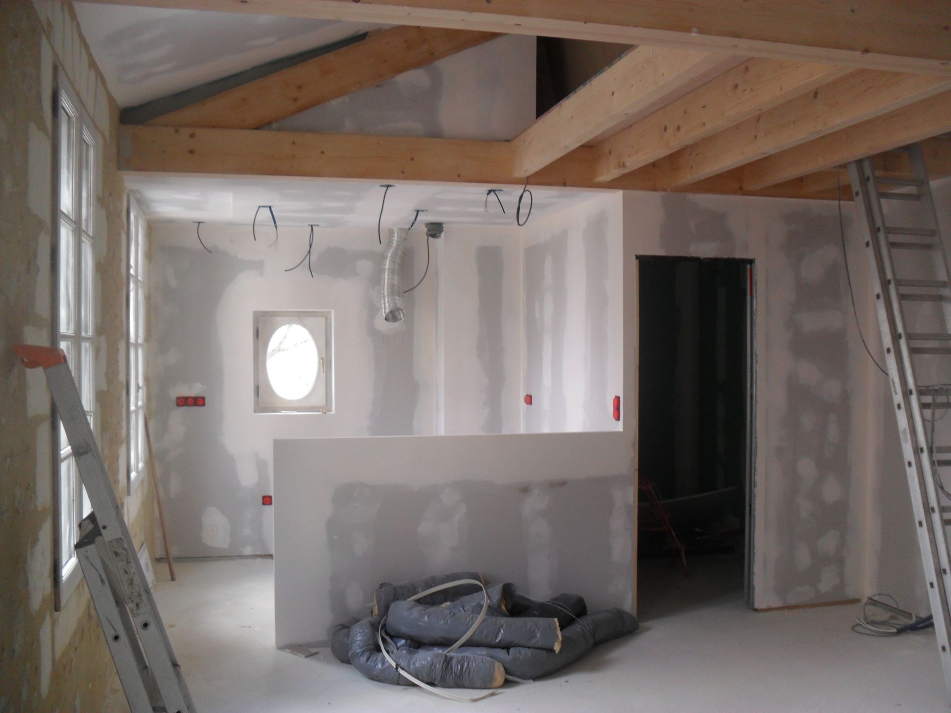 Intérieur d'une pièce en construction avec poutres apparentes, cloisons sèches, câblage électrique et une échelle.