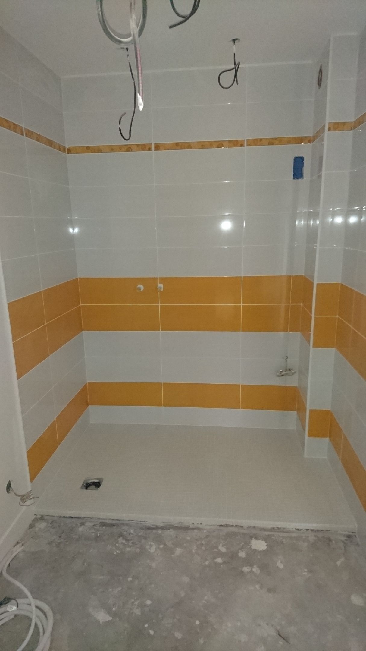 Cabine de douche avec murs carrelés blancs et jaunes, sol gris et fils électriques apparents.