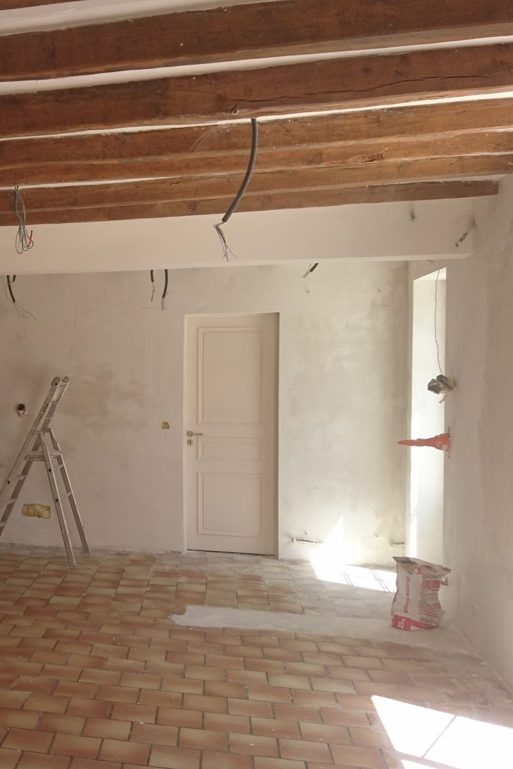 Rénovation intérieure : pièce avec poutres apparentes, murs blancs, porte et carrelage au sol. Une échelle est appuyée contre un mur.