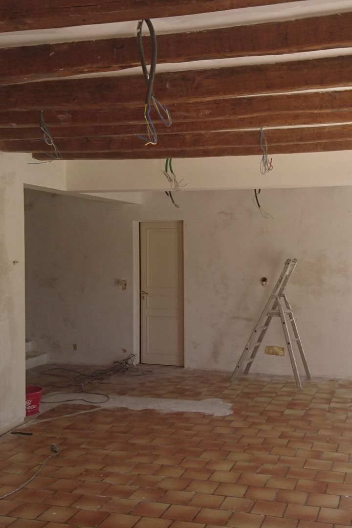 Pièce intérieure avec poutres apparentes, porte et échelle sur sol carrelé ; des câbles électriques pendent du plafond.