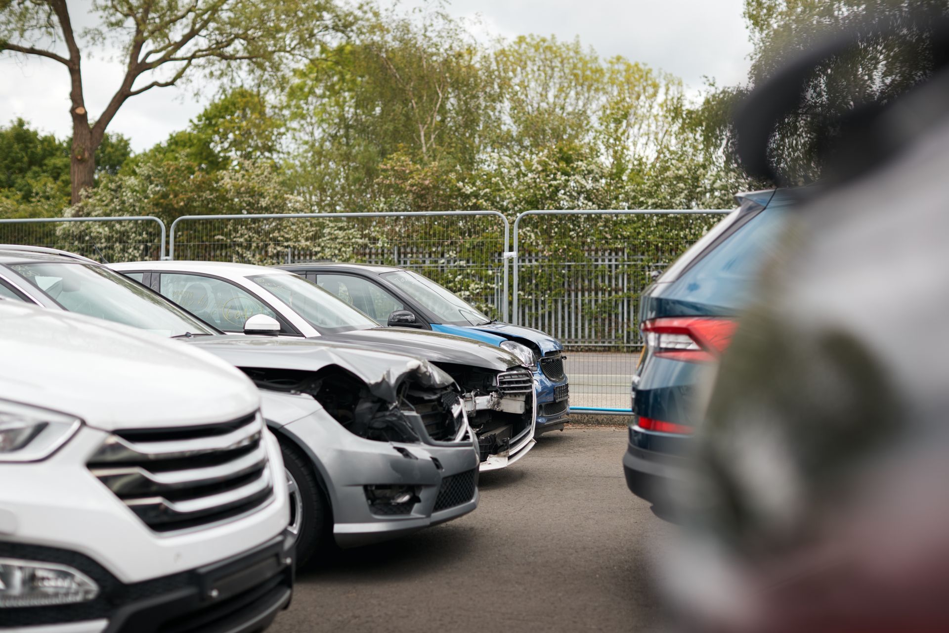 Beschädigte Autos auf einem Parkplatz, mit einem weißen SUV im Vordergrund und Bäumen hinter einem Zaun.