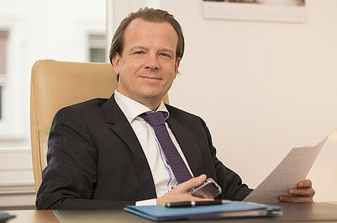 Ein Mann im Anzug sitzt an einem Schreibtisch und hält Papiere in einem Büro