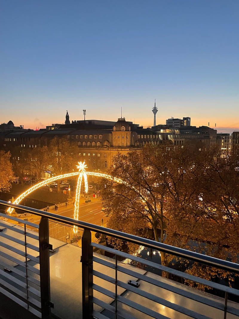 Städtische Skyline in der Abenddämmerung mit beleuchteter Brücke über einem Fluss, eingerahmt von kahlen Bäumen und einem Geländer im Vordergrund