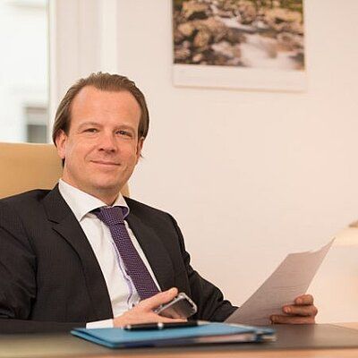 Lächelnder Mann im dunklen Anzug, der an einem Schreibtisch in einem Büro Unterlagen in der Hand hält