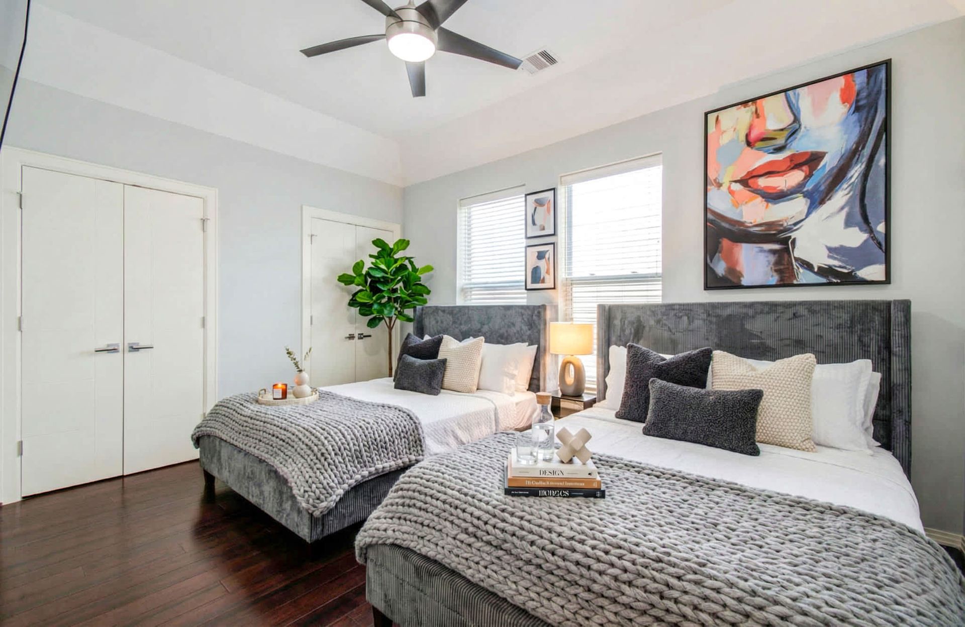Bright bedroom with two twin beds, gray bedding, dark wood floor, and colorful wall art
