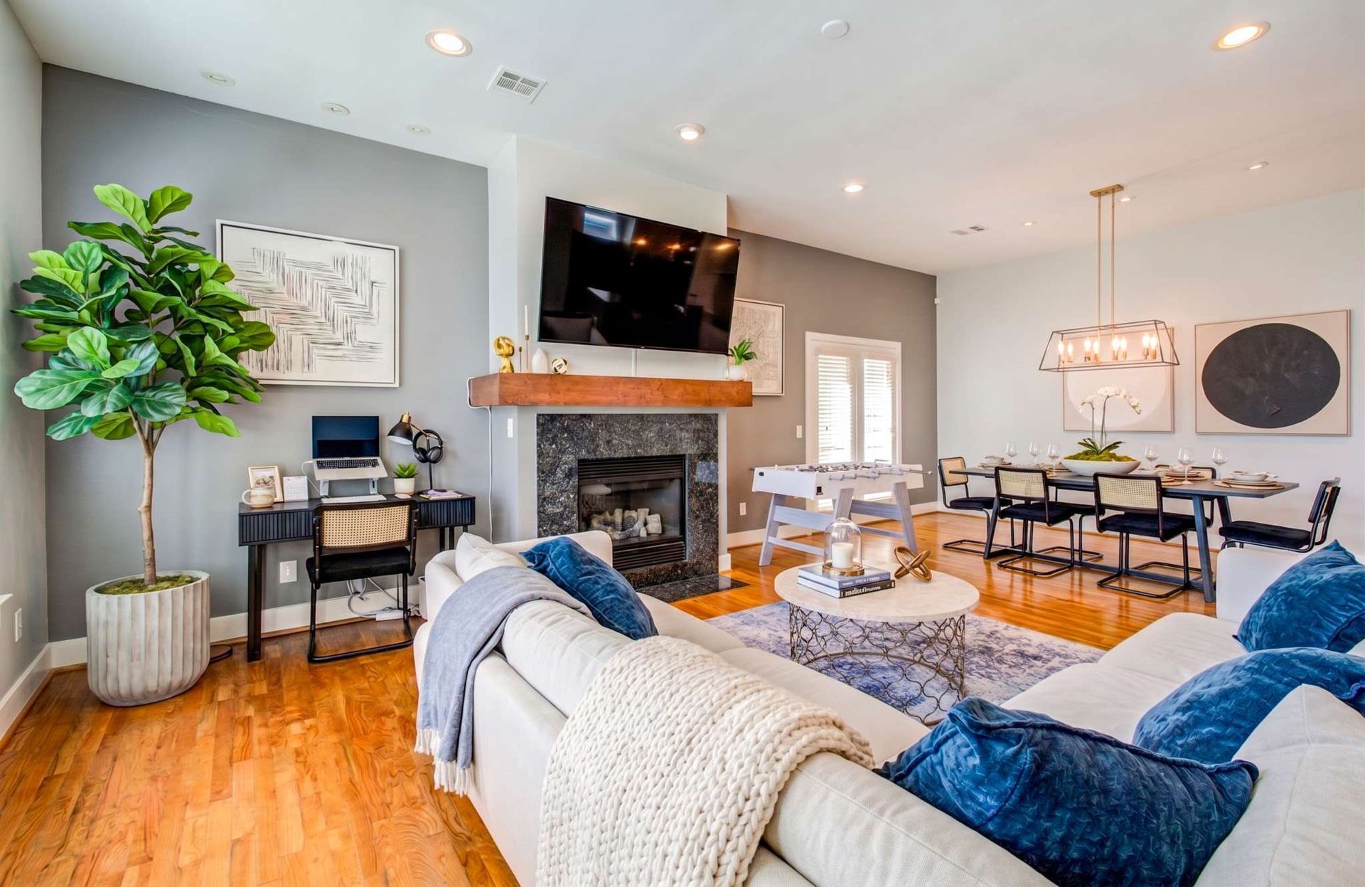 Bright living room with gray sectional, fireplace, wall-mounted TV, wood floors, and dining area.