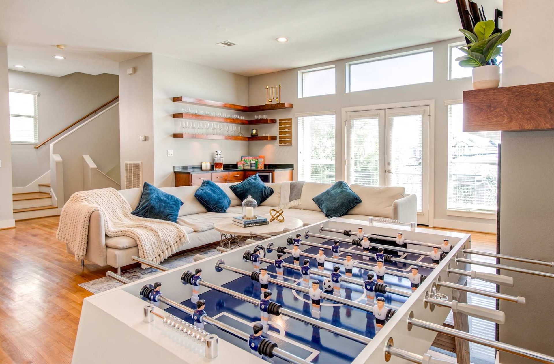 Bright living room with white sofas, blue pillows, large windows, and a foosball table in front.