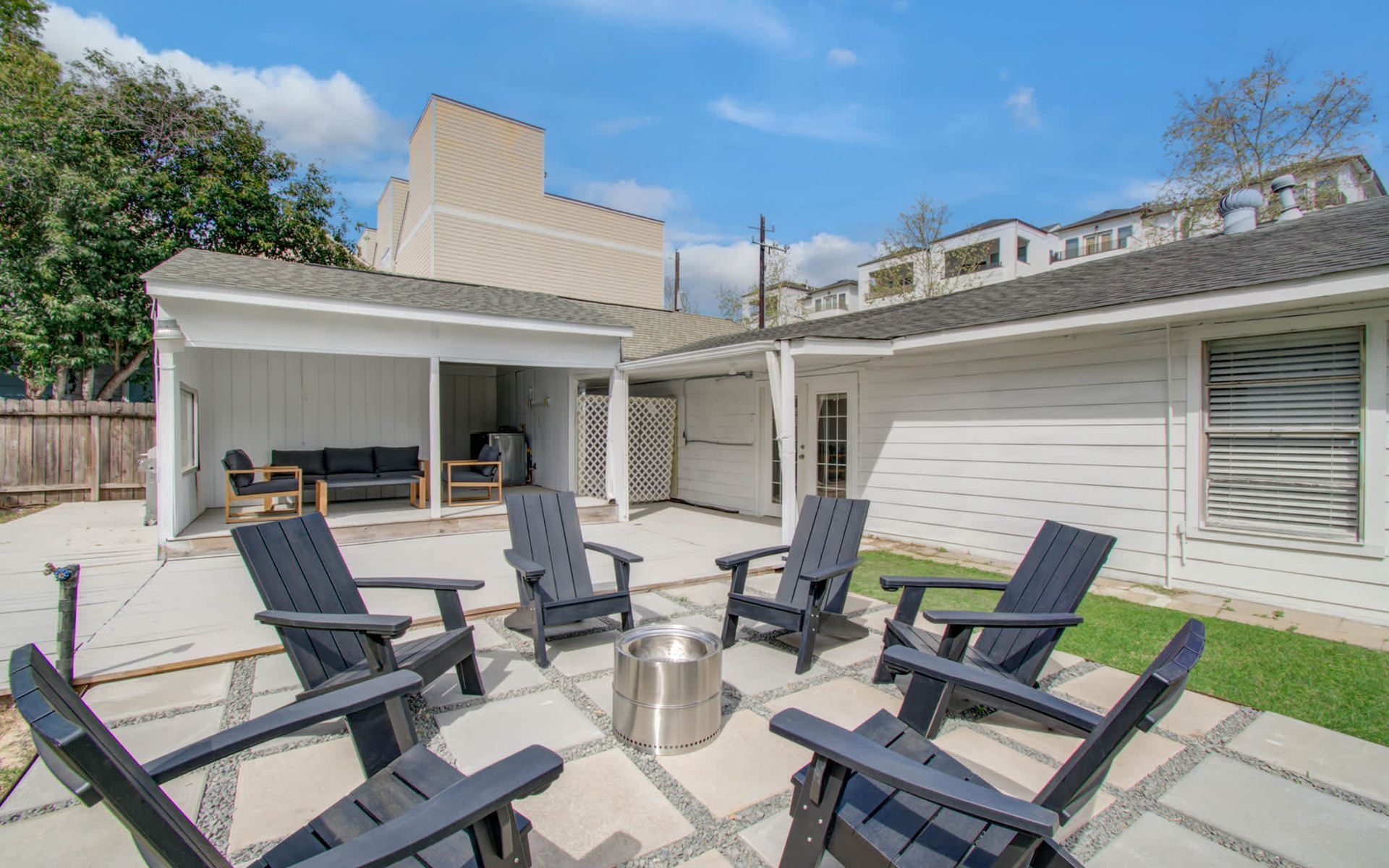 Backyard patio with six dark chairs around a fire pit beside a white house and covered seating area