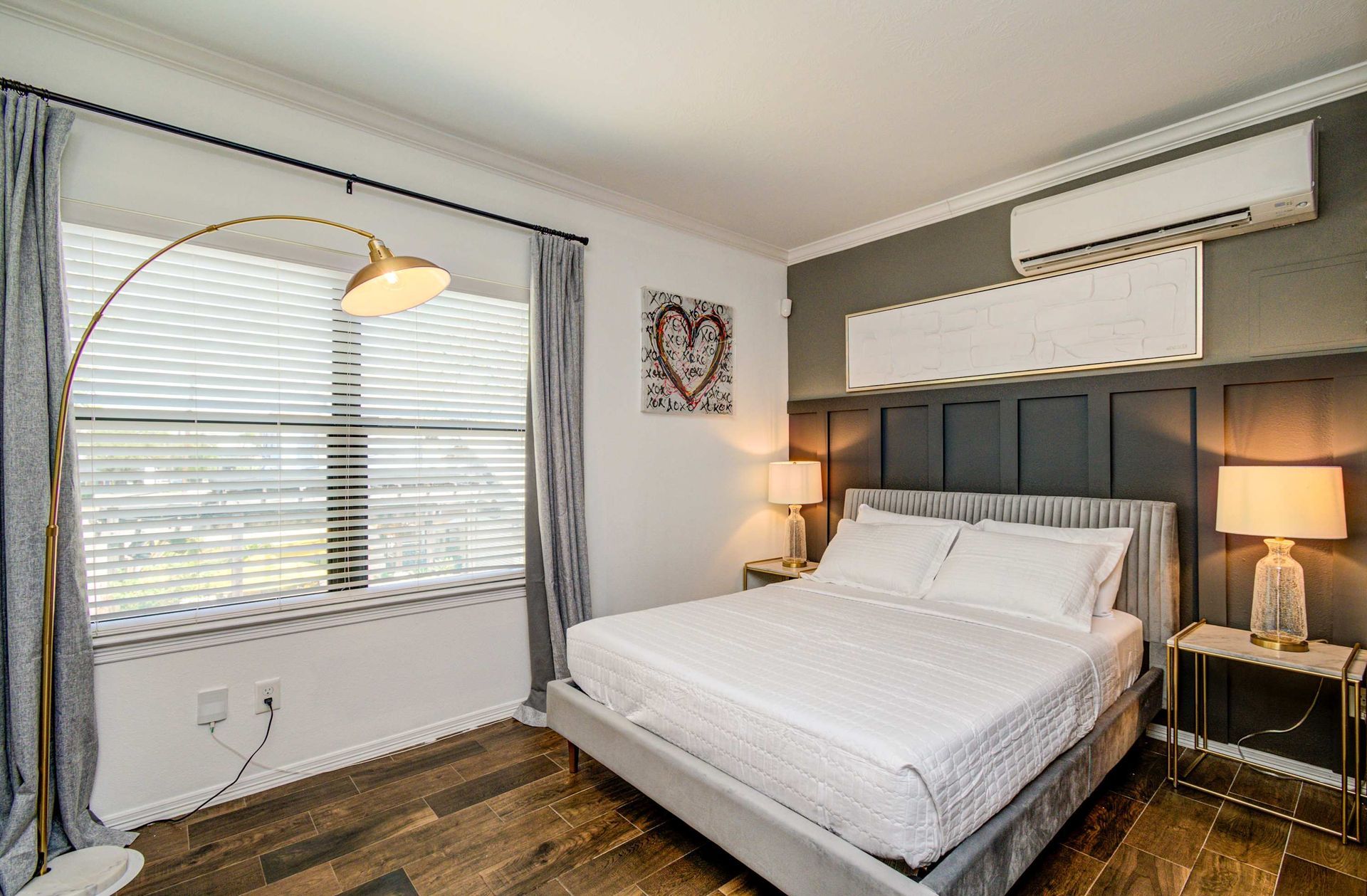 Bright bedroom with a large bed, bedside lamps, window blinds, and wall-mounted air conditioner