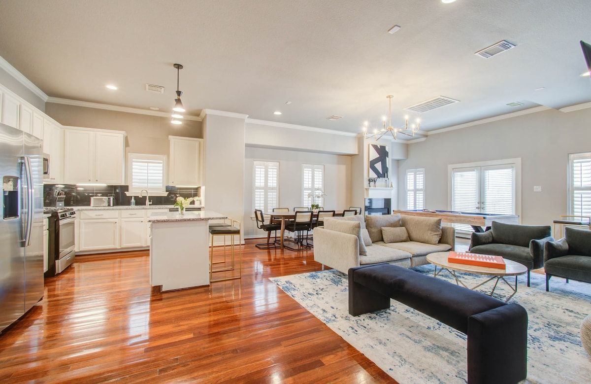 Open-concept living room and kitchen with hardwood floors, white cabinets, gray sectional, and dark ottoman.