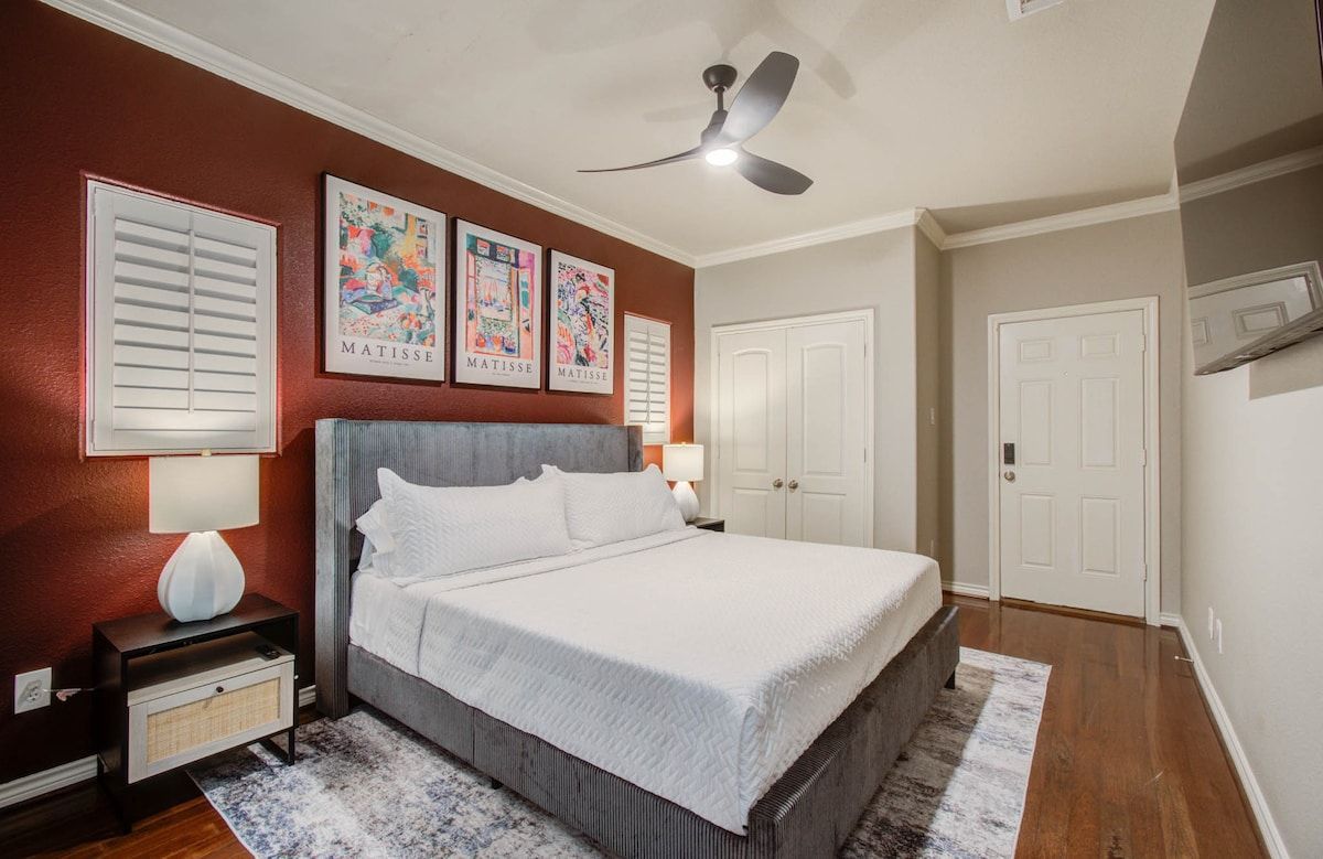 Bright bedroom with a gray bed, white walls, wood floor, and framed art above the headboard.