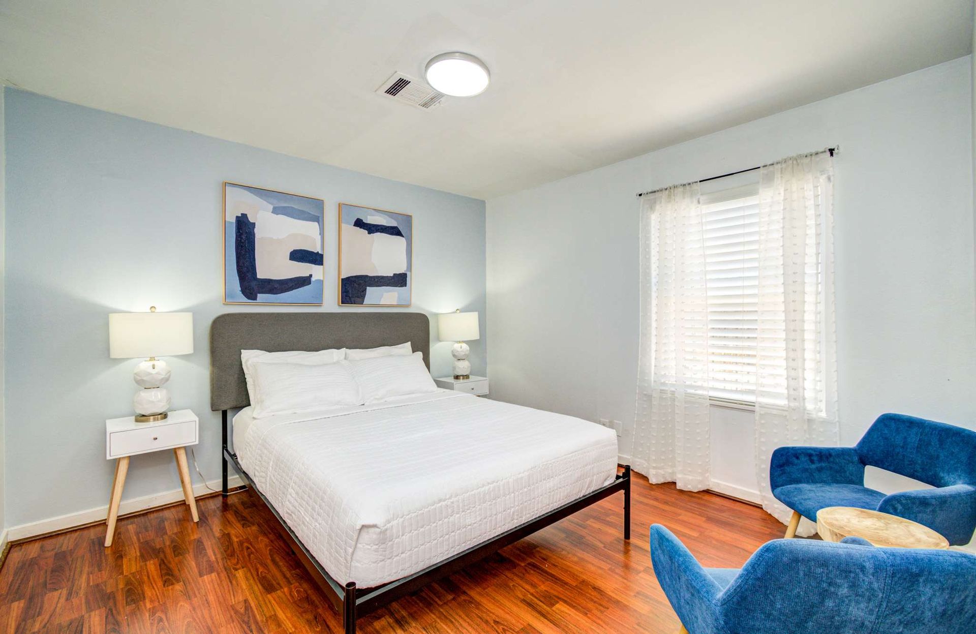 Bright bedroom with white bed, blue chairs, nightstand, and window blinds on hardwood floor