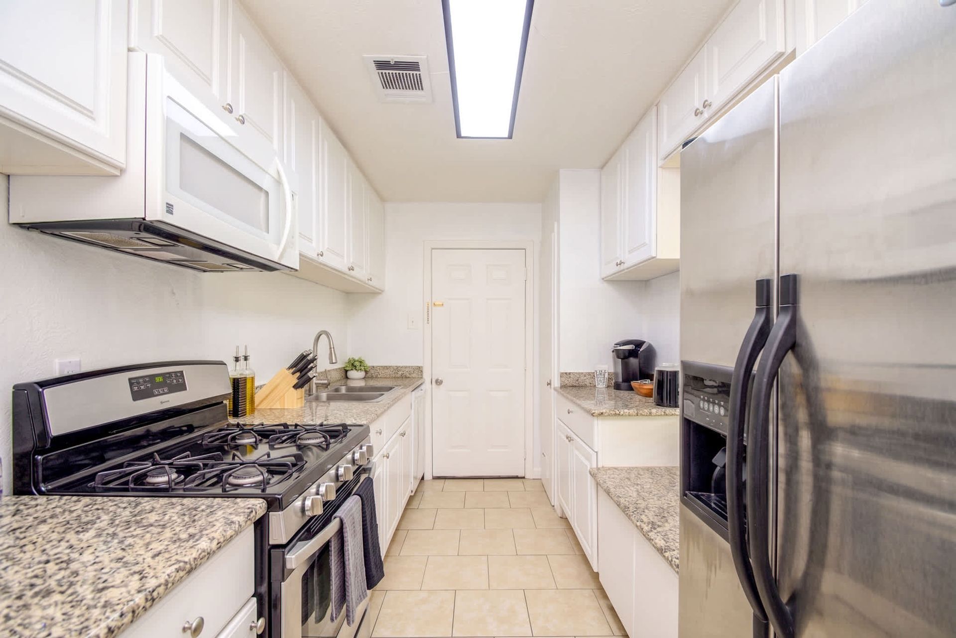 Narrow white kitchen with stainless steel fridge, gas stove, cabinets, and countertop appliances