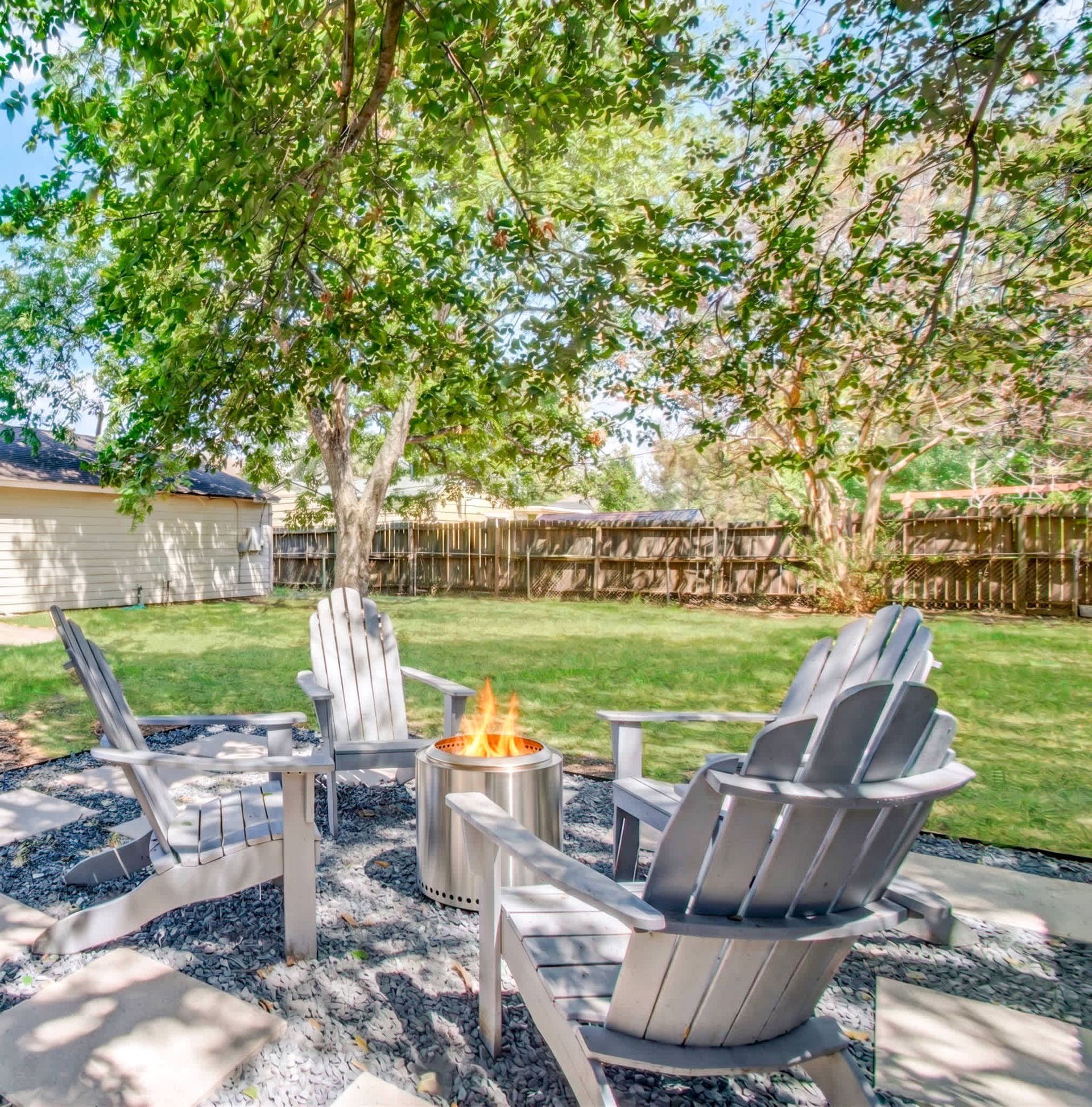 Backyard patio with gray chairs around a fire pit under a large tree, fenced lawn behind.