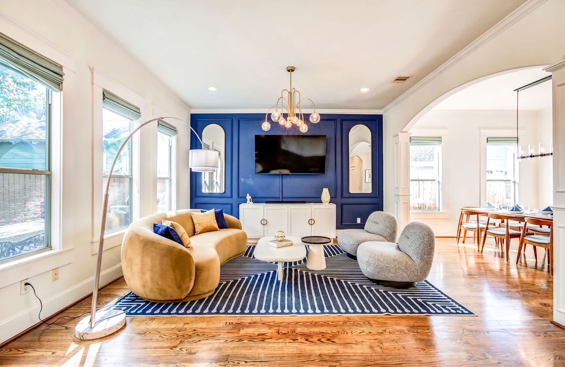 Bright living room with blue accent wall, tan chairs, TV, chandelier, and hardwood floors