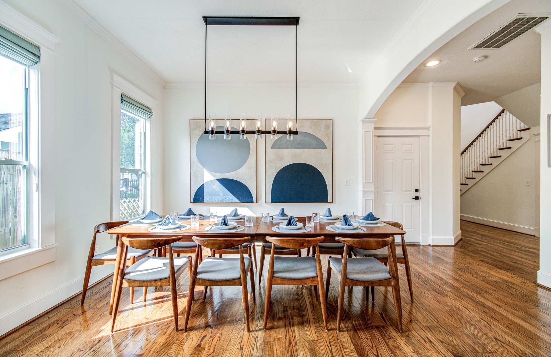Bright dining room with a long wooden table, chairs, abstract wall art, and large windows overlooking a staircase.