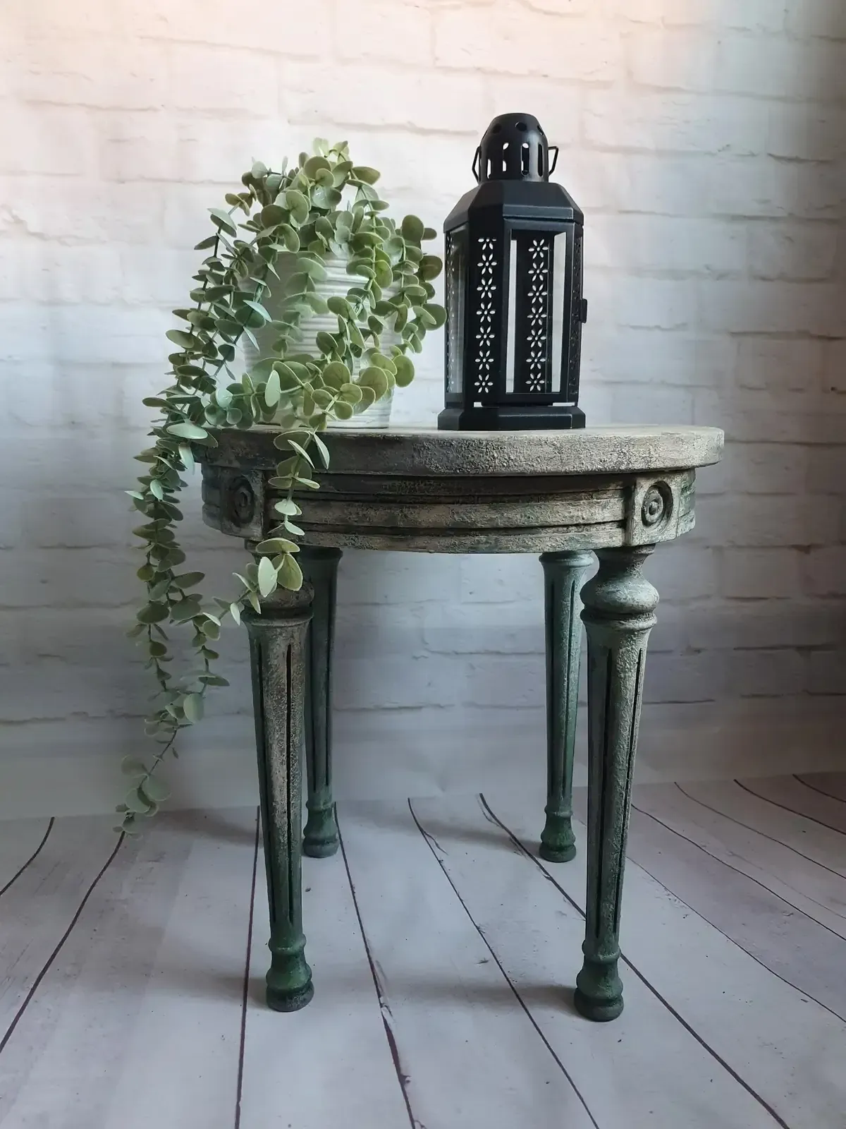 Una pequeña mesa adornada con una linterna y una planta colgante sobre un fondo de pared de ladrillo blanco.