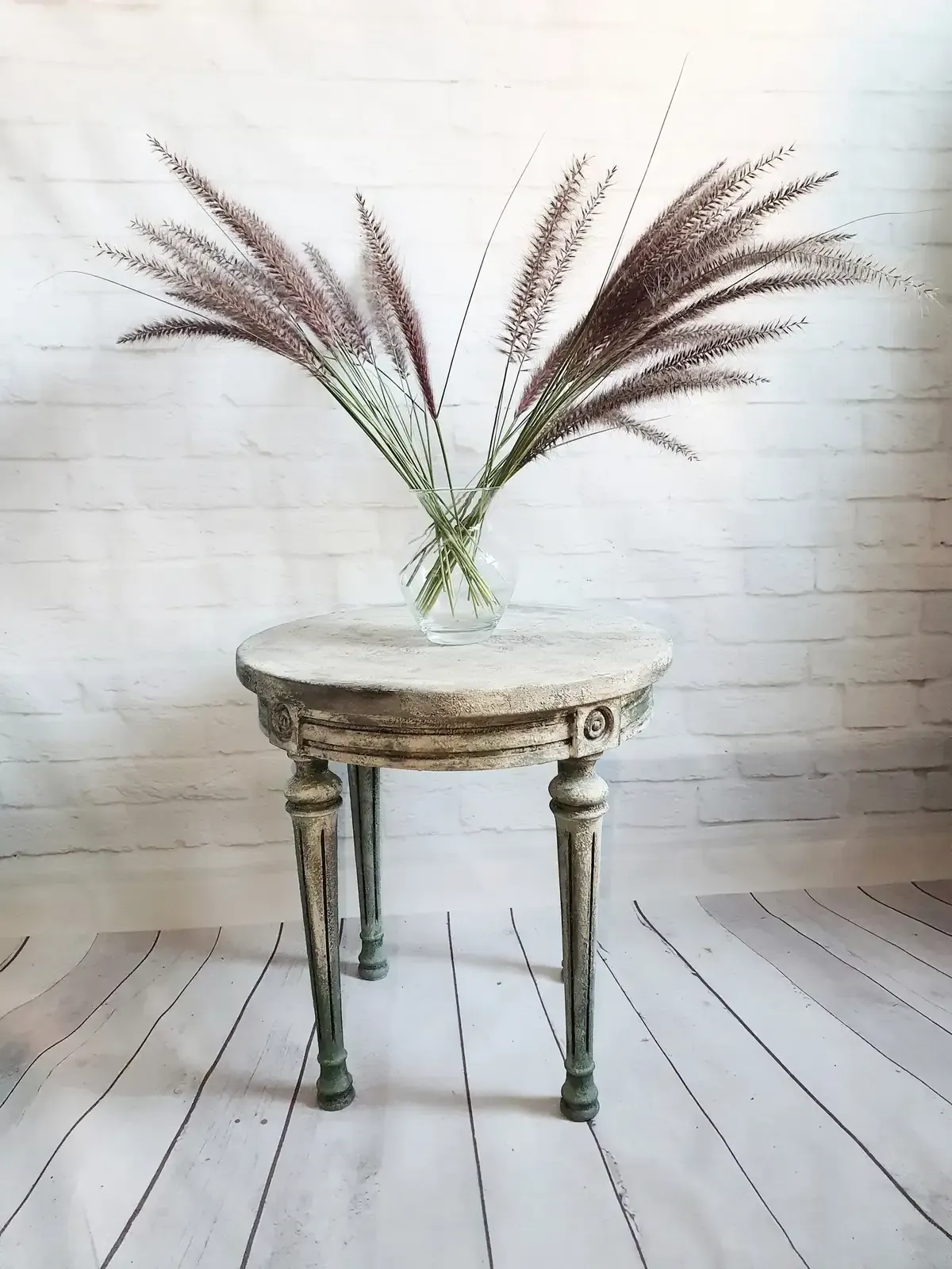 Mesa redonda desgastada con jarrón de plantas marrones y plumosas contra una pared de ladrillos blancos.