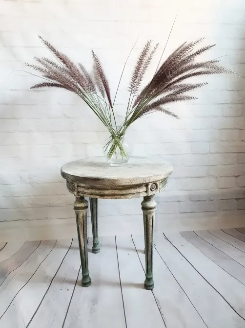 Mesa redonda desgastada con jarrón de plantas marrones y plumosas contra una pared de ladrillos blancos.