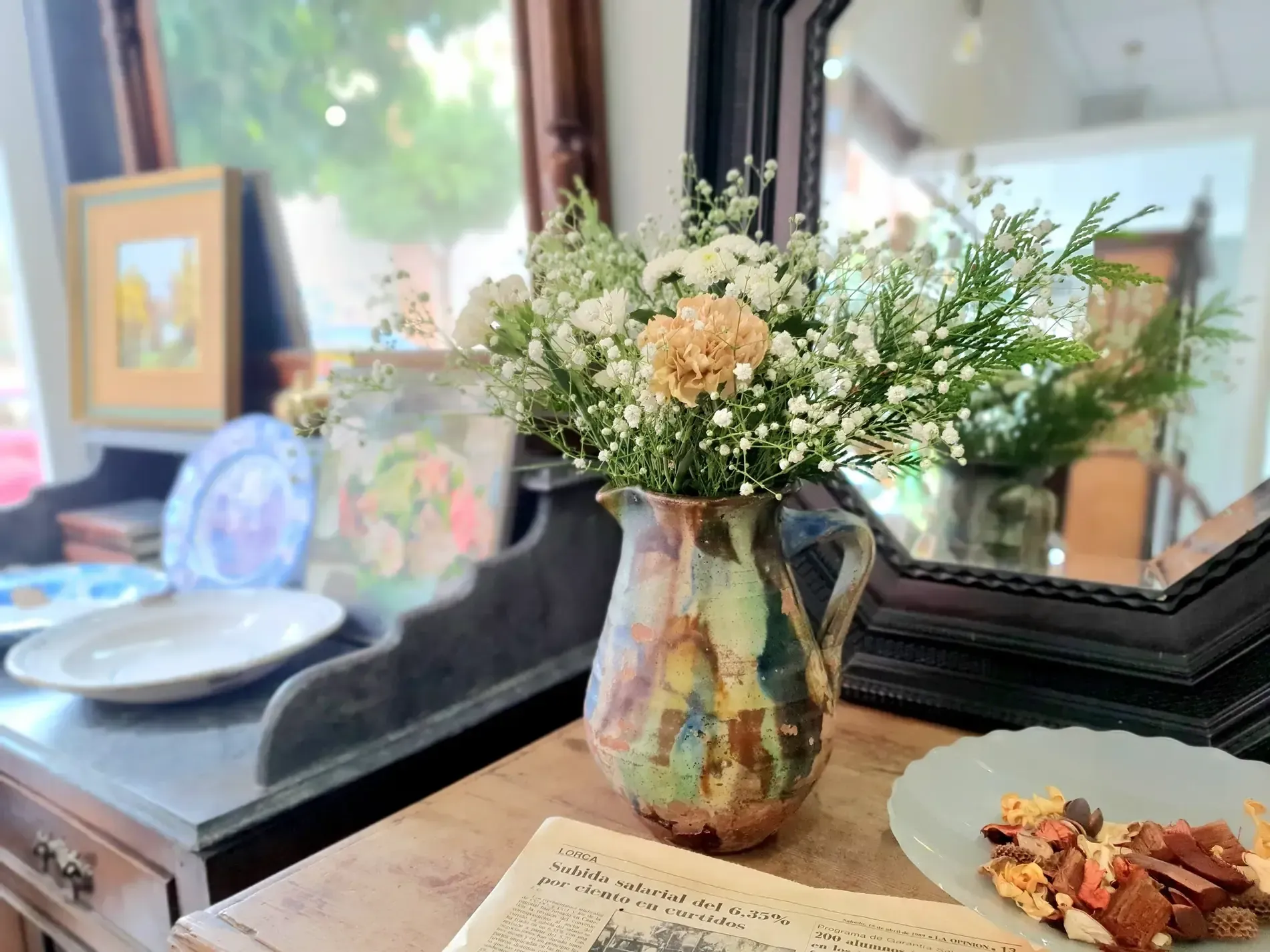 Flores en un jarrón rústico sobre una mesa, reflejadas en un espejo. También se ven platos y un periódico viejo.
