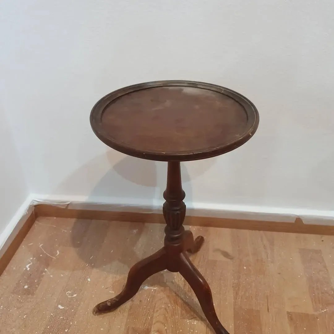 Mesa auxiliar redonda de madera con tres patas, contra pared blanca y suelo de madera.