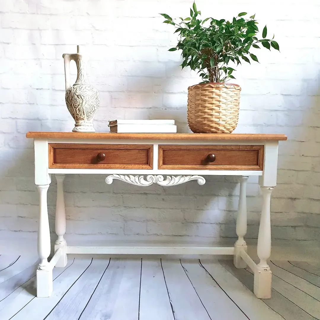 Consola de madera y color blanco con dos cajones, talla decorativa y elementos encima: jarra, libros, planta.