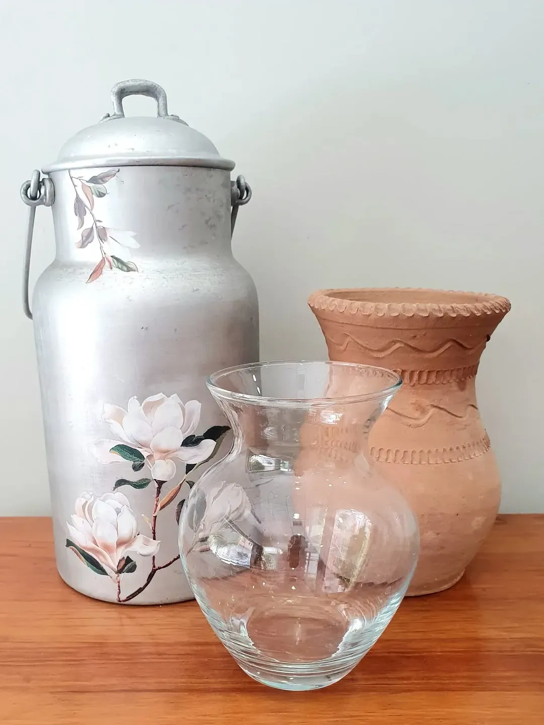 Tres jarrones sobre una superficie de madera: una lata de leche de aluminio con calcomanías florales