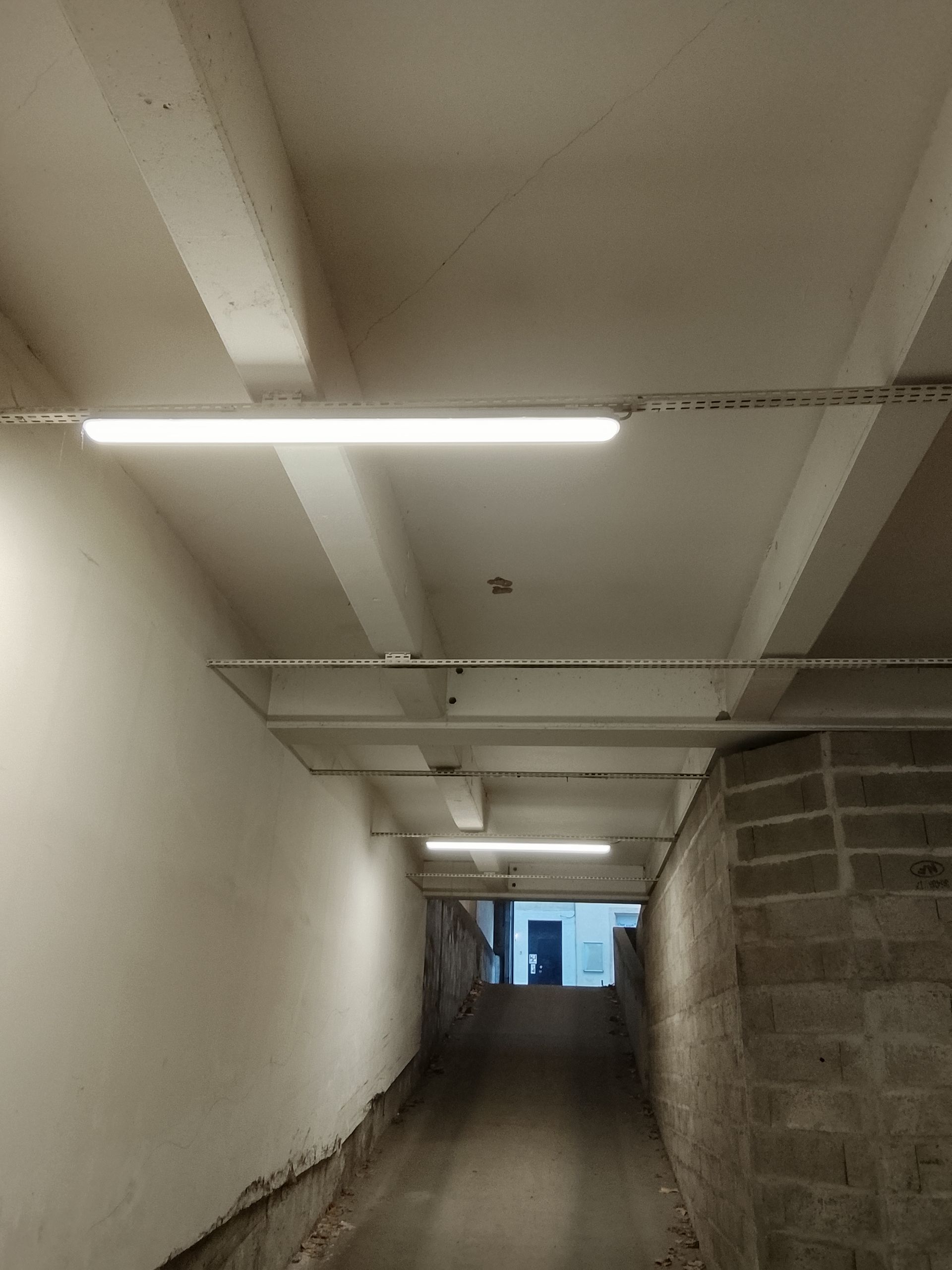 Tunnel avec éclairage fluorescent au plafond, menant à une porte visible au bout.