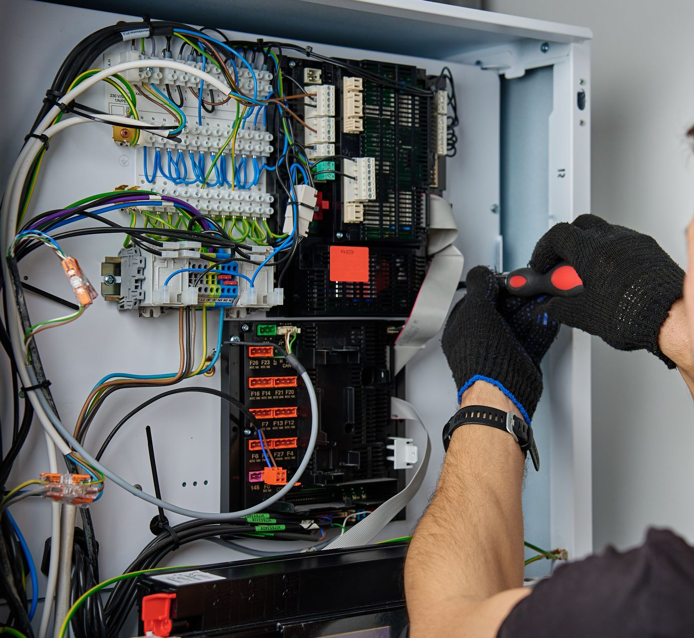 Une personne portant des gants noirs utilise un tournevis sur le câblage d'un panneau électrique à l'intérieur d'une armoire blanche.