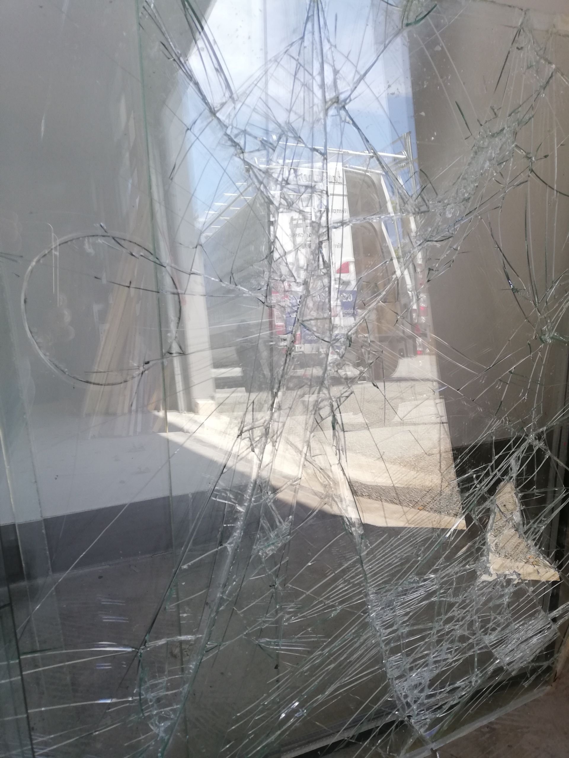 Fenêtre en verre brisé avec un réseau de fissures, révélant une scène extérieure avec des reflets de bâtiments.