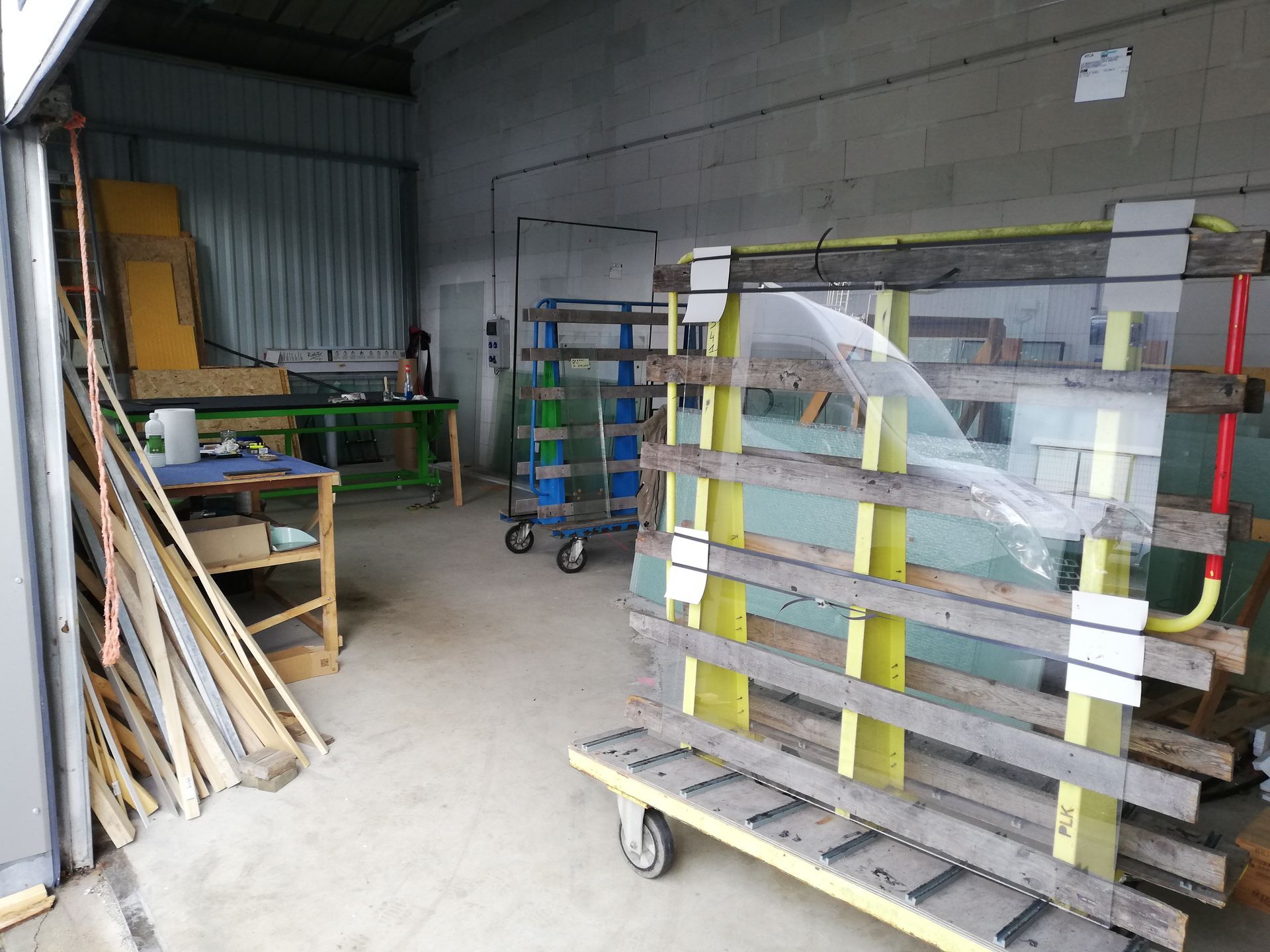 Intérieur d'entrepôt avec vitres empilées sur un chariot roulant, tables de travail et étagères de stockage.