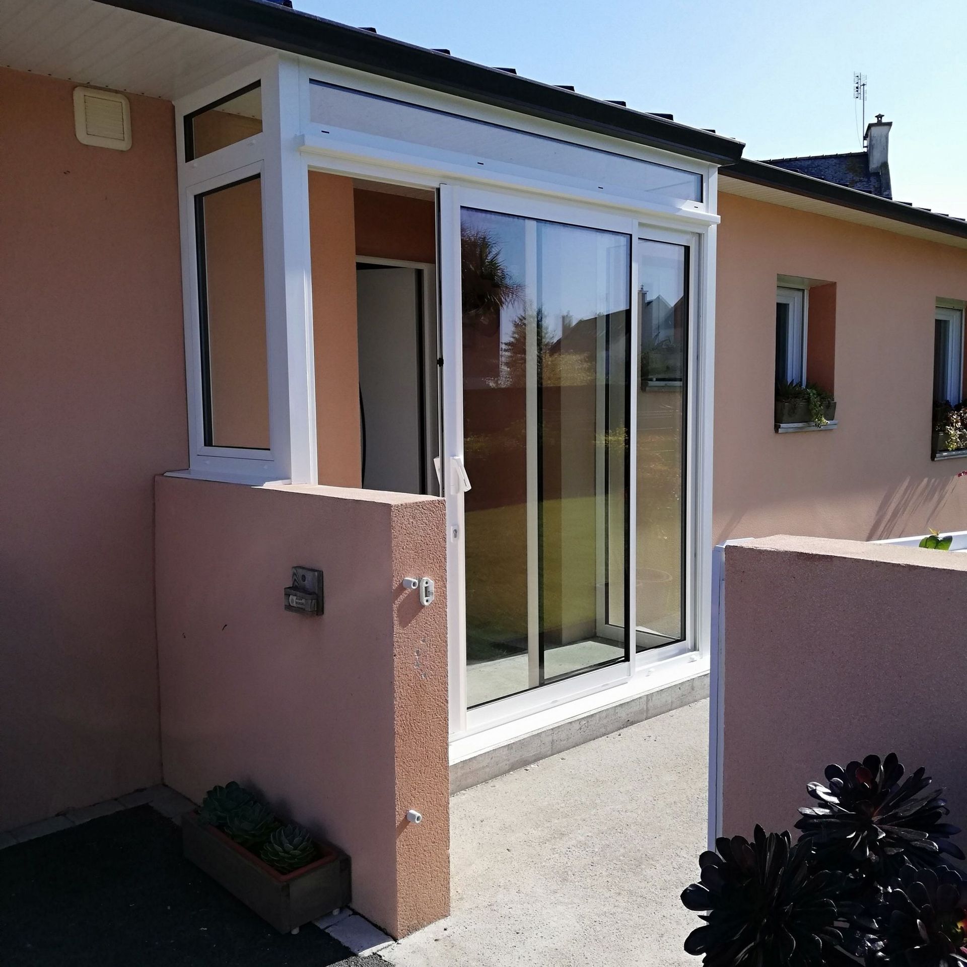 Vue extérieure d'une maison rose avec une véranda à cadre blanc, des portes coulissantes en verre et un petit mur en béton.