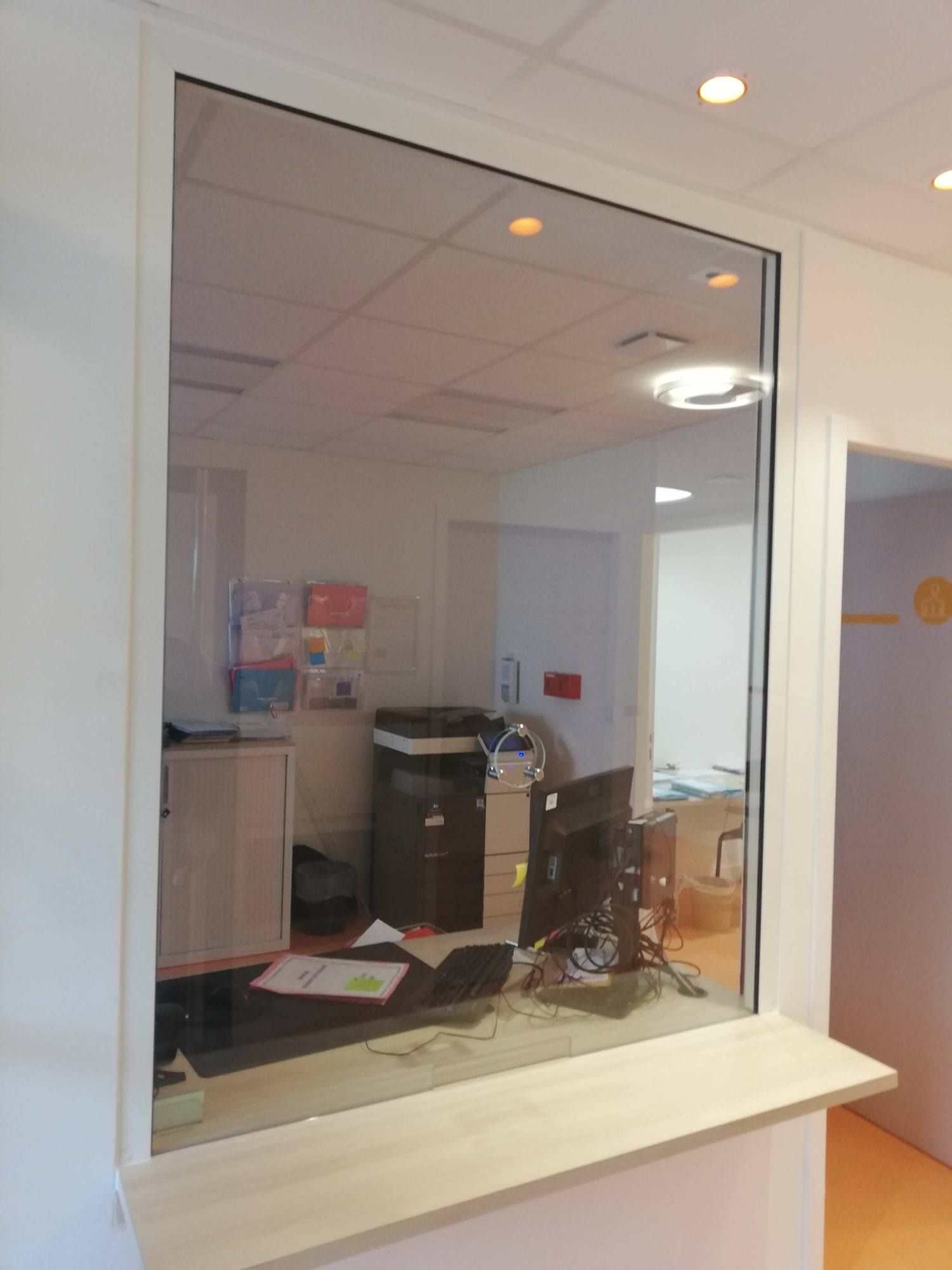 Fenêtre de réception avec vitre transparente ; vue sur les bureaux. Comptoir en bois clair.