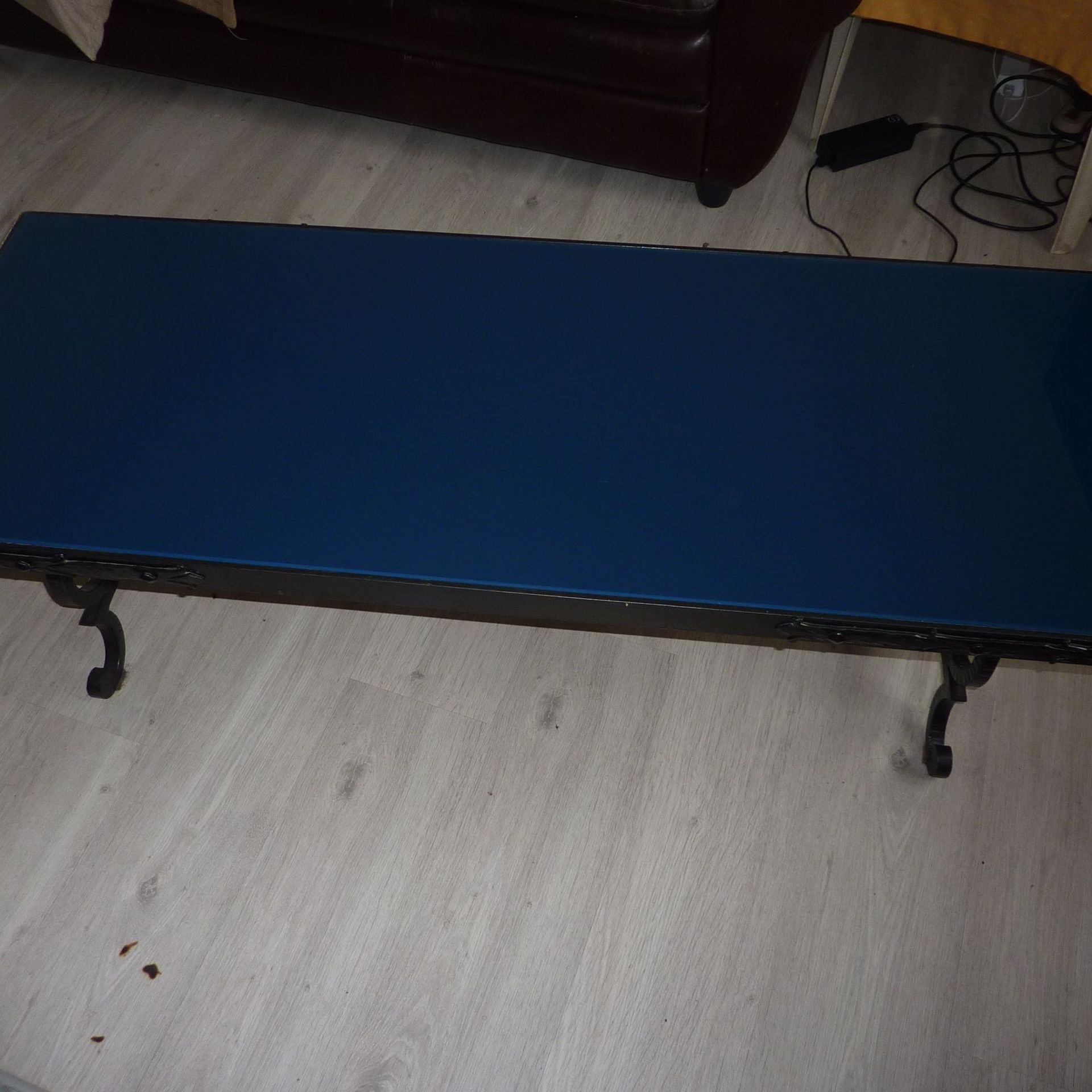 Table basse rectangulaire bleue avec pieds noirs ornés. Sol gris et meubles sombres en arrière-plan.
