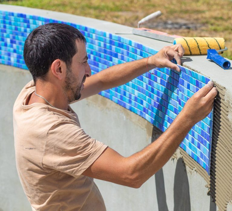 Un trabajador presiona cuidadosamente una lámina de mosaico azul sobre la pared de hormigón de una piscina con adhesivo.