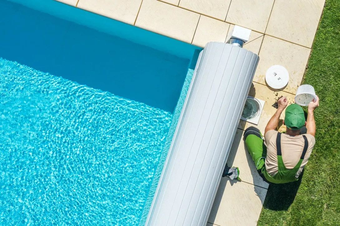 Un técnico con ropa de trabajo verde está arrodillado junto a una piscina, realizando el mantenimiento de un equipo.