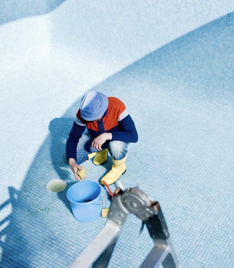 Una persona con un sombrero azul y botas amarillas limpia una piscina seca y vacía con una esponja y un cubo azul.