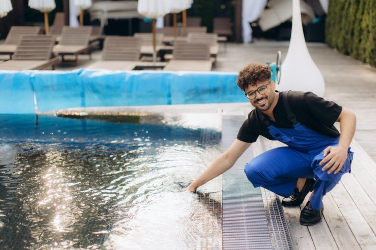 Un técnico con mono azul está arrodillado junto a una piscina, comprobando la calidad del agua cerca del borde de la terraza.