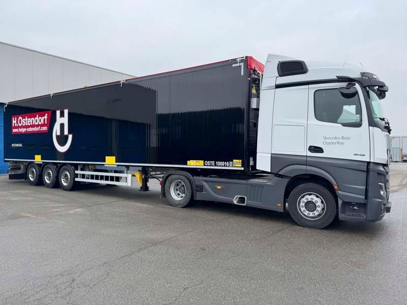Draußen parkt ein weißer Mercedes-Sattelzug mit schwarzem Anhänger. Auf dem Anhänger sind ein Logo und eine Website zu sehen.