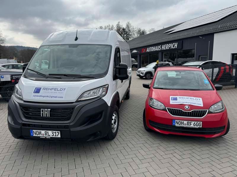 Weißer Fiat-Transporter und roter Skoda auf einem gepflasterten Parkplatz vor einem Autohaus geparkt, bewölkter Himmel.