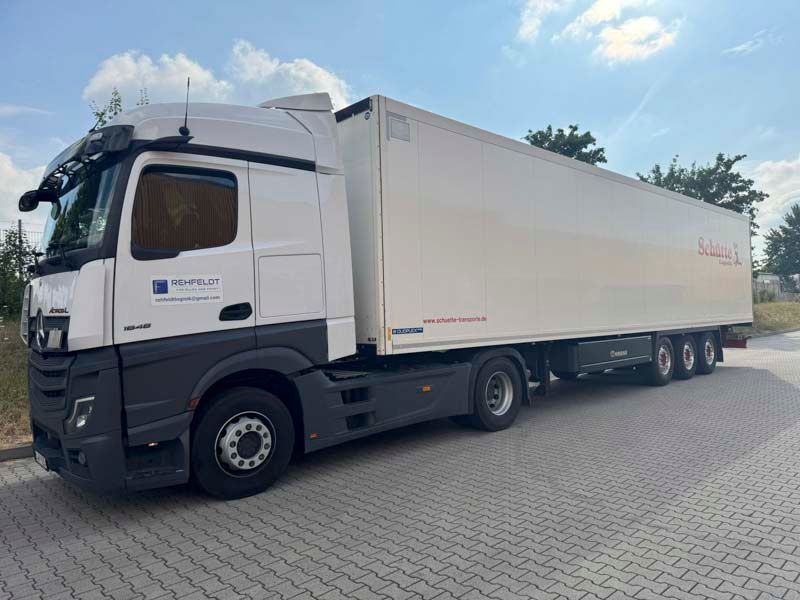Weißer Sattelschlepper mit Anhänger auf einem gepflasterten Parkplatz unter blauem Himmel. Der LKW hat schwarze und graue Akzente und einige Firmendetails.