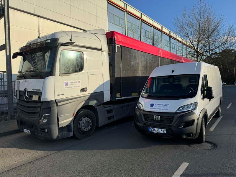 LKW und weißer Lieferwagen parken nebeneinander, beide mit demselben Logo. Gebäude im Hintergrund.