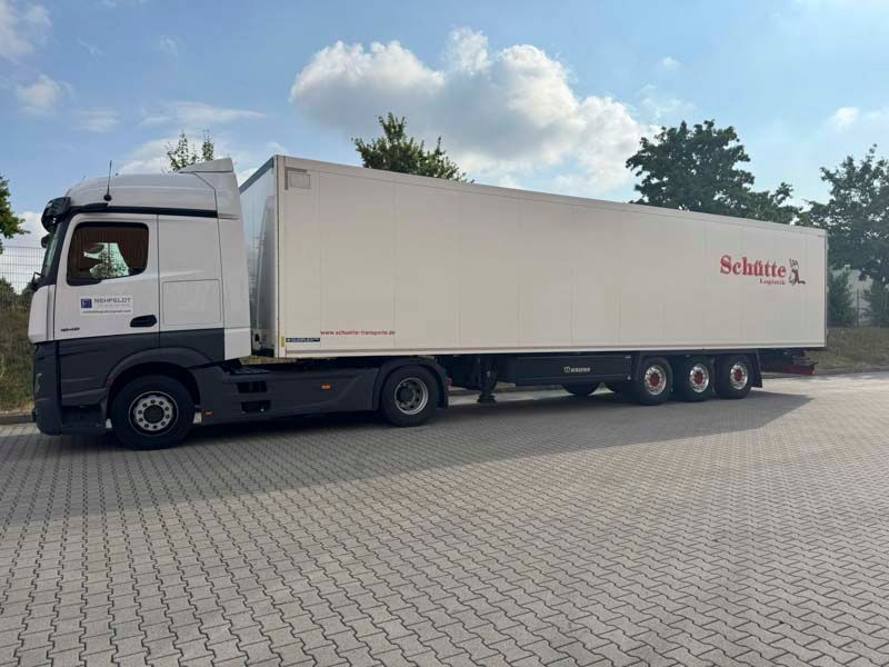 Weißer Sattelschlepper mit Anhänger auf einem gepflasterten Parkplatz geparkt. Bäume und blauer Himmel im Hintergrund.