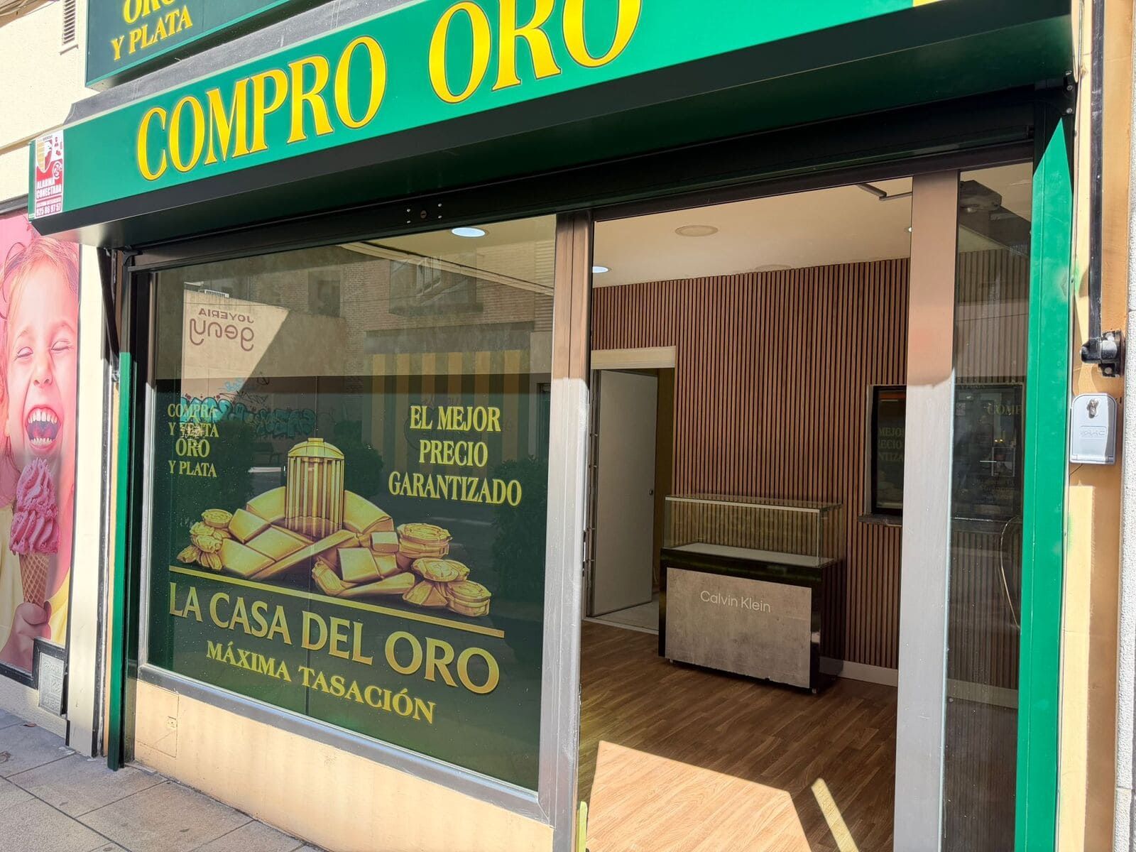 Fachada de la tienda de compra de oro “Compro Oro” con letrero verde y escaparate en una calle soleada.