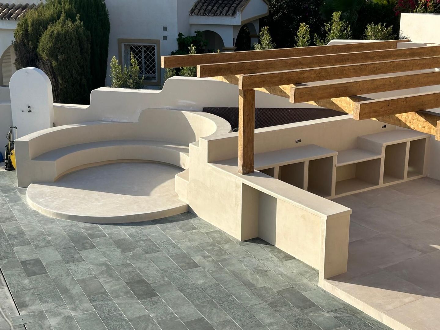 Patio moderno con asientos curvos blancos integrados, pérgola de madera y adoquines de piedra gris bañado por la luz del sol.