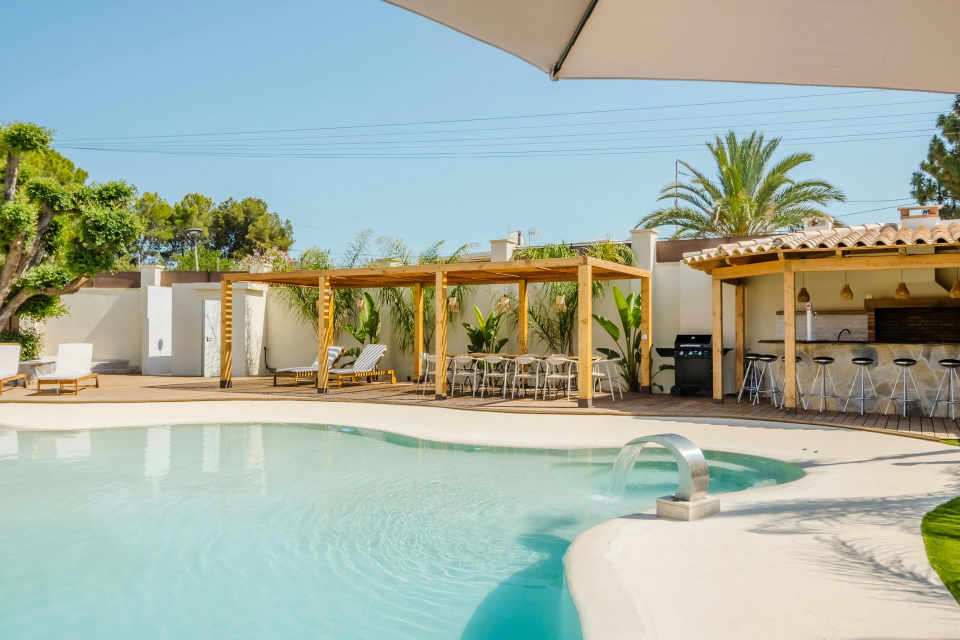 Piscina soleada del complejo con pérgola, palmeras y zona de bar junto a aguas cristalinas.