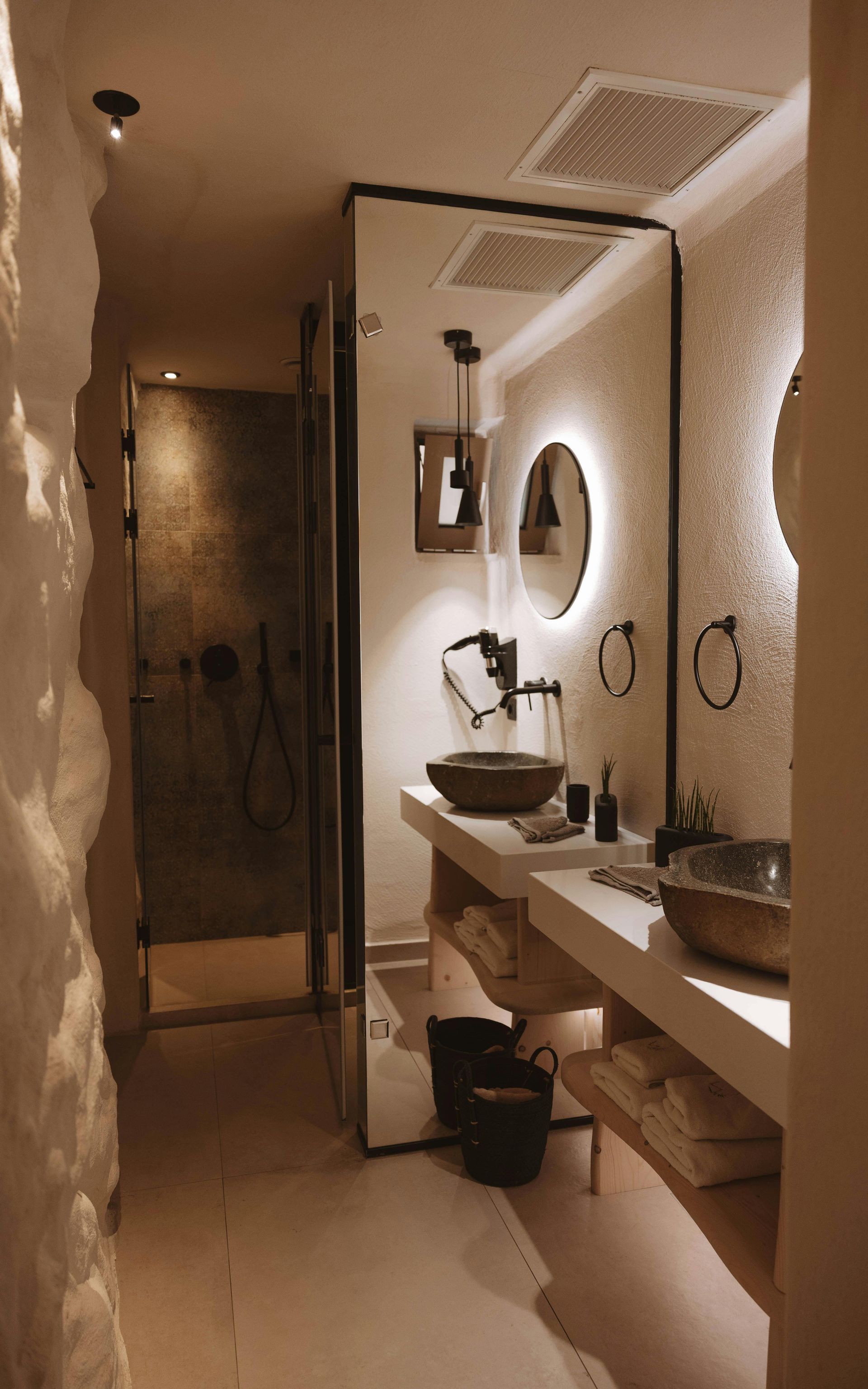 Baño moderno de color beige con tocador largo, espejo redondo, lavabo, ducha e iluminación cálida.